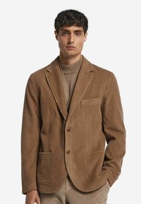 Brauner Baumwollcord-Blazer mit einem Reverskragen, zwei Fronttaschen, einem Knopfverschluss und gerippter Textur. Über einem beigen Rollkragenpullover getragen.