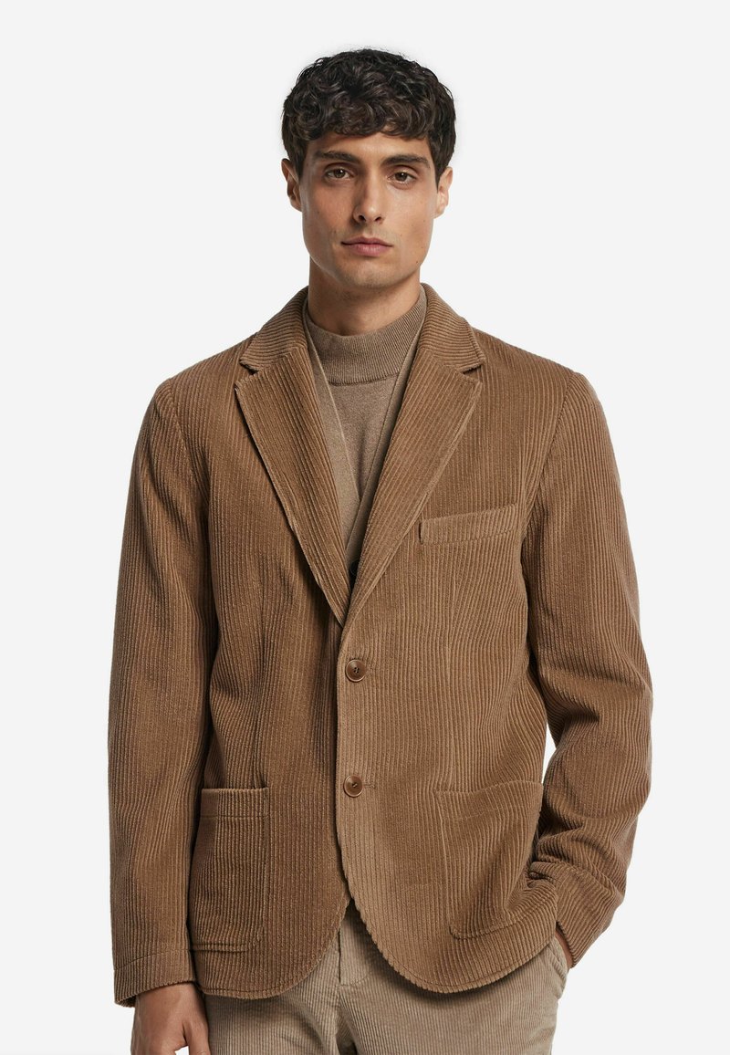 Brauner Baumwollcord-Blazer mit einem Reverskragen, zwei Fronttaschen, einem Knopfverschluss und gerippter Textur. Über einem beigen Rollkragenpullover getragen.