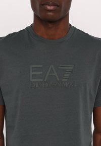 Siva kratka majica s kratkimi rokavi, izdelana iz mehkega materiala, s teksturiranim logotipom EA7 in blagovno znamko "EMPORIO ARMANI" na sprednji strani.