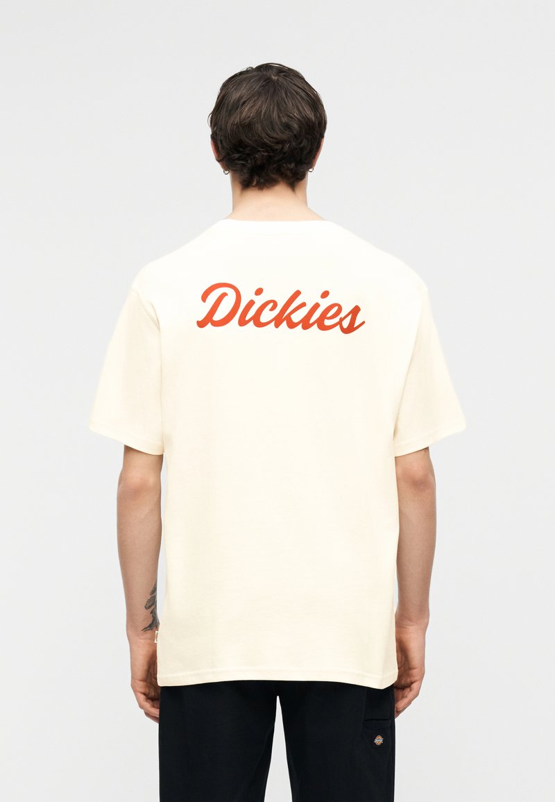 Persona con cabello corto y oscuro que lleva una camiseta crema con el texto naranja "Dickies" en la parte posterior y pantalones negros, de pie frente a un fondo liso.