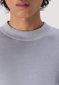 Lys grå sweatshirt med ribbet rund hals, glat tekstur og subtile stikninger, vist fra brystet og op.