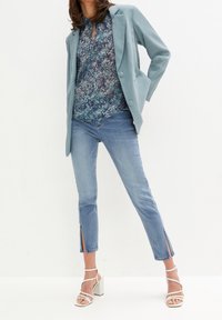 Blazer léger bleu sarcelle porté sur un chemisier bleu à motifs, associé à un jean en denim clair avec des fentes sur les côtés, et des sandales blanches à talons blocs.