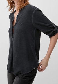 Donna che indossa una blusa grigia scura a maniche lunghe con le maniche arrotolate e pantaloni scuri, in piedi contro uno sfondo chiaro e semplice.