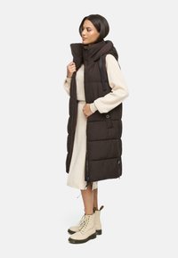 Veste matelassée marron longue et sans manches avec un col haut, des poches latérales et une fermeture éclair. Portée sur une robe de couleur claire.