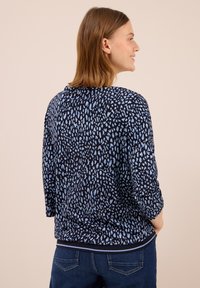 Frau mit schulterlangem Haar, die ein langärmeliges, navyblaues Oberteil mit hellblauen abstrakten Punkten und dunkelblauen Jeans trägt und seitlich blickt.