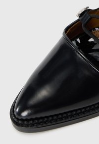 Schwarzer Lacklederschuh mit spitz zulaufender Zehe, geflochtenen Details entlang der Sohle und einem schwarzen Riemen mit einem dekorativen silbernen Akzent.