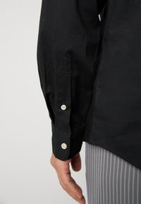 Schwarz langärmliges Hemd mit knöpfbaren Manschetten, glattem Stoff, seitlichen Schlitzen und geradem Saum. Gepunktete, gestreifte Hose ist sichtbar.