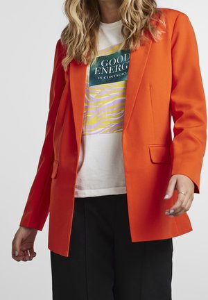 Femme portant un blazer orange vif sur un t-shirt blanc avec le texte « Good Energy is Contagious » et un pantalon noir, debout devant un fond uni.