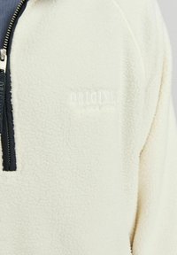 Cremfarbener Fleece-Pullover mit hohem Kragen, halbseitigem Reißverschluss und besticktem "ORIGINA"-Logo. Strukturierte Oberfläche, Reißverschluss in Kontrastfarbe.