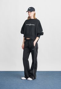 Μαύρο κοντό φούτερ με το λογότυπο "NIKE SHOX", συνδυασμένο με μαύρο παντελόνι που έχει πλευρικές λευκές λωρίδες, και μαύρο καπέλο. Λευκά παπούτσια ολοκληρώνουν την εμφάνιση.