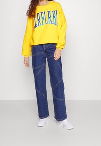 Sudadera amarilla con letras azules, combinada con pantalones de mezclilla anchos azules que presentan detalles de costura en diagonal. Zapatillas blancas completan el look.