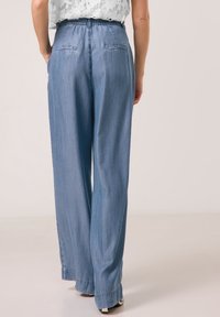 Kobieta nosząca jasnoniebieskie dżinsy z wysokim stanem i szerokimi nogawkami z rozcięciami na końcach oraz białą koronkową bluzkę, stojąca na jednolitym jasnym tle.