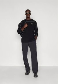 Sort langermet skjorte med hvite kontrastsømmer, logopatch; grå bukser; svart ryggsekk; hvite solbriller; svarte joggesko.