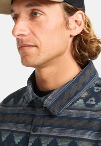 Patterned jacket with a blue base, featuring geometric designs in teal and brown. Black snap buttons and a collar with contrasting stripes.