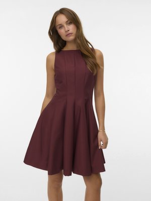 Kobieta o długich brązowych włosach, ubrana w bezrękawową bordową sukienkę o linii A, stojąca na jednolitym białym tle.