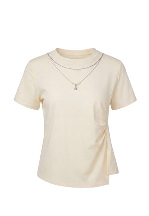 T-shirt color crema a maniche corte con leggero drappeggio laterale, abbinata a due collane d’argento sovrapposte, una con ciondolo a stella.