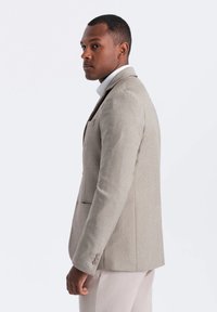 Blazer beige ajusté avec un revers cranté, une poche unique et des poignets à trois boutons. Texture lisse, coupe structurée.