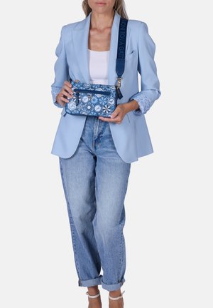 Woman wearing light blue blazer and jeans holds blue patterned crossbody bag with wide strap over shoulder.