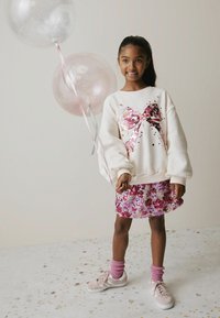 Hvid sweatshirt med et stort grafisk bue i pink perler; pink og lilla nederdel med perler; pink sokker; hvide sneakers; holder klare balloner.