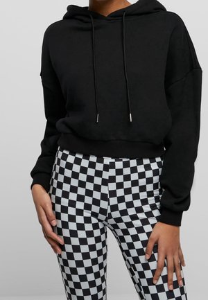 Person iført en sort cropped hoodie og sort-hvide ternede bukser med høj talje, stående med hænderne afslappet ned langs siderne.