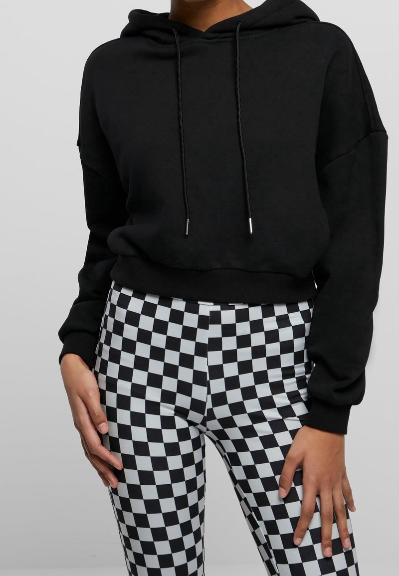 Personne portant un sweat à capuche court noir et un pantalon à carreaux noir et blanc taille haute, debout les mains détendues le long du corps.