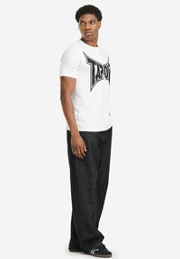 Weißes T-Shirt mit kurzen Ärmeln und schwarzem Logodesign. Kombiniert mit dunkelblauen Jeans und schwarzen Sneakern. Schlicht, locker sitzend.