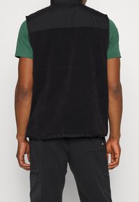 Černá fleecová vesta s vysokým límcem, texturovaným předním dílem a hladkým zadním dílem. Má nastavitelné zapínání a rovný střih. Vhodná pro vrstvení.