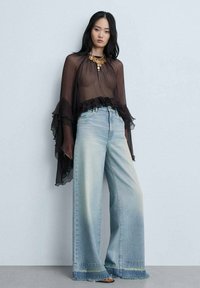 Black sheer top with ruffled edges, paired with light blue wide-leg jeans featuring frayed hems. Simple brown sandals complete the look.