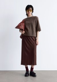 Frau in einem übergroßen braunen Pullover, brauem Midikleid, burgunderfarbenen Socken mit Plateauslippern, gelb getönten Sonnenbrille, die eine große geflochtene Clutch hält.