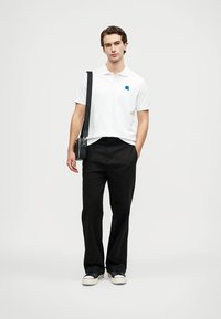 Biela polokošeľa s modrým logom, čierne široké nohavice a tmavé tenisky. Modelka stojí s čiernou ramennou taškou. Hladký, ležérny vzhľad.