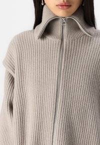 Pull en maille côtelée beige avec un col montant et une fermeture éclair métallique. Présente une coupe décontractée et une texture douce.