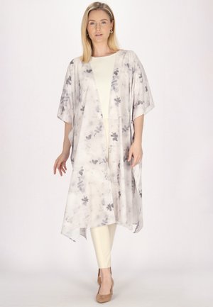 Blondýna nosící dlouhé, vzdušné světlé kimono se šedými květinovými vzory, krémové kalhoty a světle hnědé podpatky, stojící na jednobarevném pozadí.