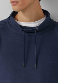 Felpa con cappuccio blu navy con colletto alto, coulisse e trama a coste sulle cuciture delle spalle; indossata sopra una t-shirt bianca.