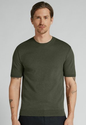 Mężczyzna o jasnej karnacji i średnio brązowych włosach ma na sobie gładki oliwkowozielony sweter z krótkim rękawem, stoi na jednolitym jasnoszarym tle.