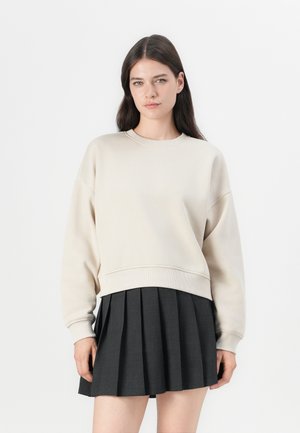 Beige genser med rund hals og senkede skuldre, kombinert med et svart plissert skjørt. Mykt stoff med ribbestrikkede mansjetter og nederkant.