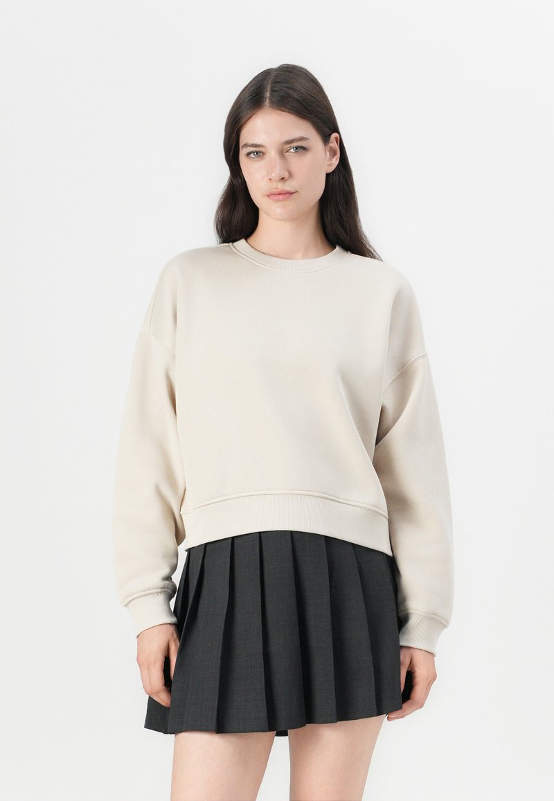 Beige genser med rund hals og senkede skuldre, kombinert med et svart plissert skjørt. Mykt stoff med ribbestrikkede mansjetter og nederkant.