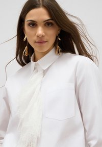 Chemise blanche à col avec un panneau en dentelle au niveau du cou. Elle possède une poche plaquée et des manches longues. Boucles d'oreilles décoratives en or. Texture lisse.