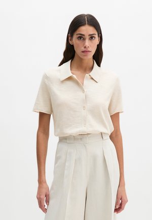 Kobieta o długich ciemnych włosach, ubrana w kremową, krótką koszulę z guzikami, wpuszczoną w wysokowalne, jasnobeżowe spodnie z plisami.