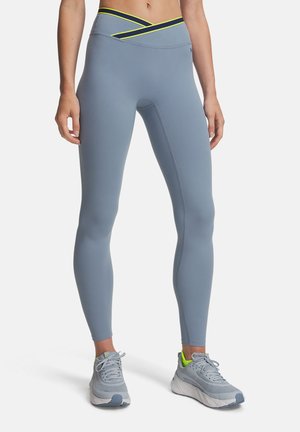 Woman wearing blue-gray high-waist leggings with yellow and black waistband detail and gray athletic shoes standing on white background.