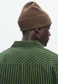 Grünes Hemd mit Knopfleiste und vertikalen hellgrünen Streifen; weiche Textur. Braune gerippte Strickmütze. Ansicht von hinten.