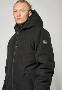 Giacca nera con cappuccio dal tocco morbido, dotata di zip, tasca sul petto e toppa con logo sulla manica. Design casual per abbigliamento urbano.