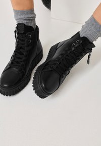 Crne visoke tenisice od glatke kože, s teksturiranom gumenom potplatom, vezanjem sprijeda i patentnim zatvaračem sa strane.