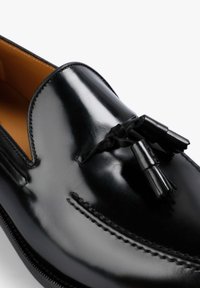 Mocasín de cuero negro con un acabado brillante, que presenta tres borlas decorativas en la parte frontal y costuras detalladas a lo largo de los bordes.