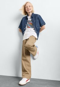 Camicia in denim con bottoni, t-shirt bianca grafica, pantaloni beige e sneaker bianche con accenti rossi e blu. Bambino in posa contro un muro.