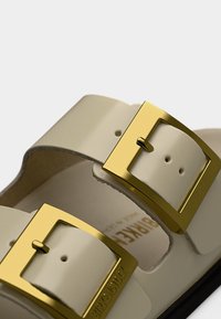 Μπεζ σανδάλια με δύο χρυσά αγκράφια, κατασκευασμένα από λείο συνθετικό υλικό. Διαθέτουν διαμορφωμένο πέλμα και επίπεδη εξωτερική σόλα.