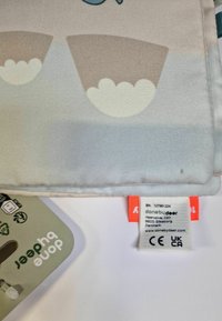 Manta de bebé de tejido suave en colores pasteles claros, con patrones de nubes. Etiqueta adjunta con detalles de la marca e instrucciones de cuidado.