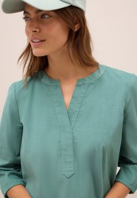 Giovane donna che indossa una blusa turchese con scollatura a fessura e un cappellino chiaro, che guarda leggermente verso sinistra su uno sfondo semplice.