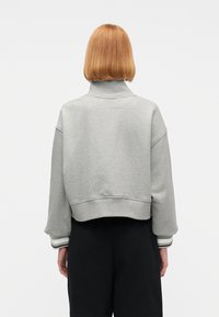 Άτομο με κόκκινα μαλλιά μήκους έως τους ώμους που φοράει γκρι κοντή μπλούζα με ζιβάγκο και μαύρο παντελόνι, γυρισμένο με την πλάτη, πάνω σε λευκό φόντο.