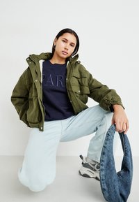 Casaco acolchoado verde-oliva com fecho e capuz forrado, usado sobre um sweatshirt azul-marinho. Jeans azul-claro e ténis cinzentos completam o visual.