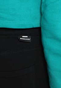 Tela turquesa de una camisa parcialmente superpuesta sobre pantalones negros. Una etiqueta en los pantalones dice "DRDENIM JEANMAKERS." Presenta costuras oscuras y un bolsillo.